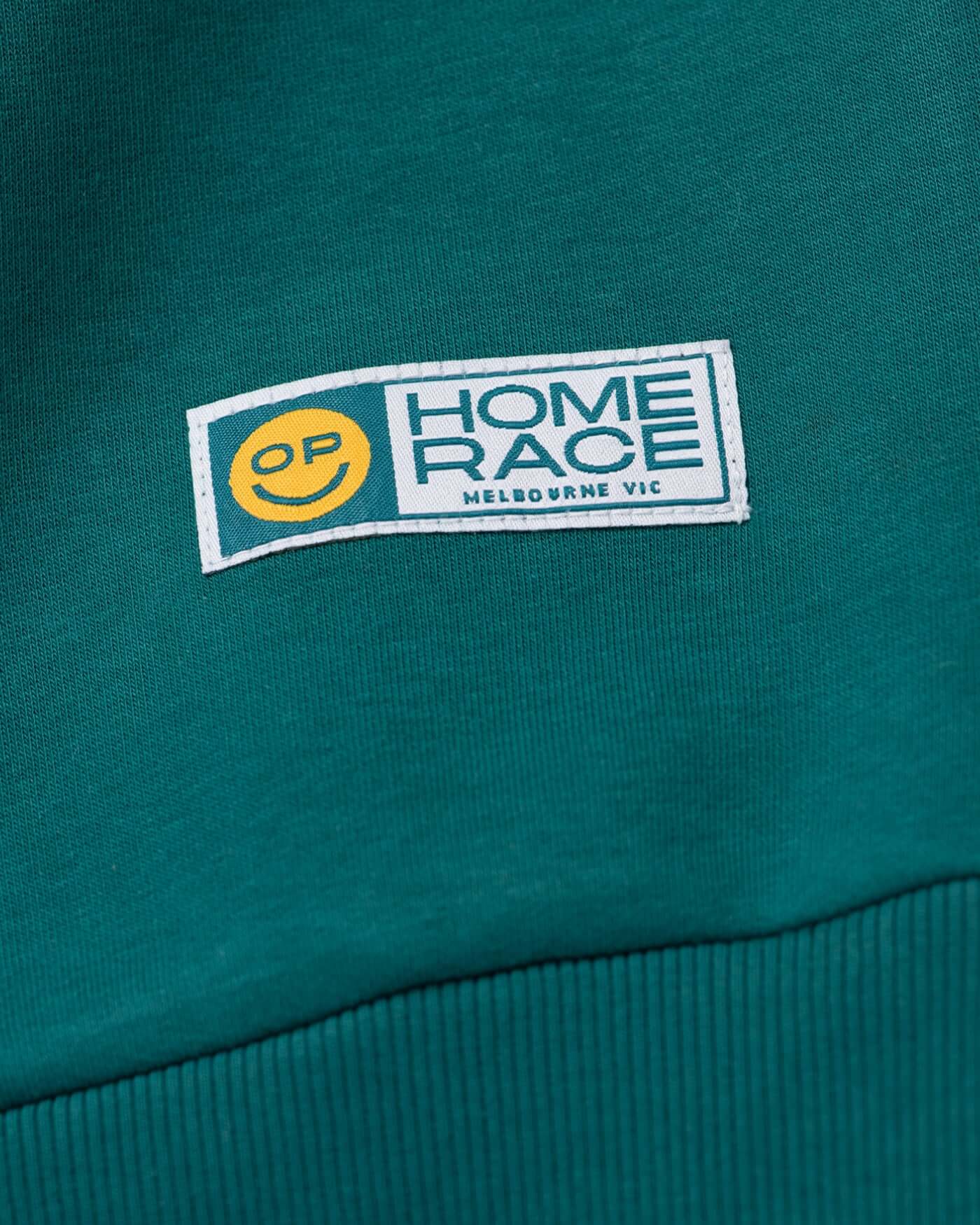 Close-up of the label on the Home Race '26 Sweater featuring the OP smiley logo and Melbourne VIC.