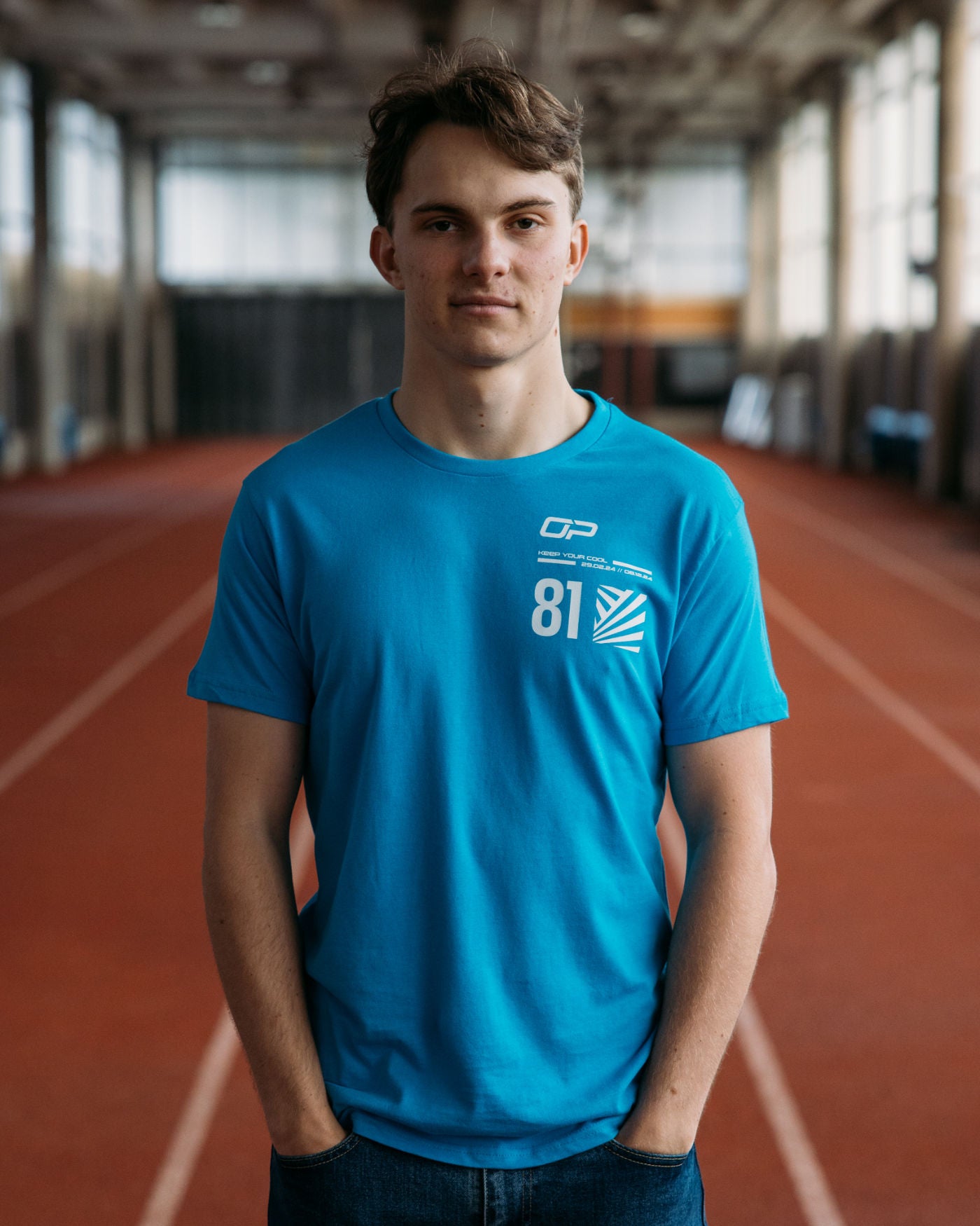 Man wearing Keep Your Cool T-Shirt in Aqua featuring OP logo and dazzle effect, standing on an indoor track.