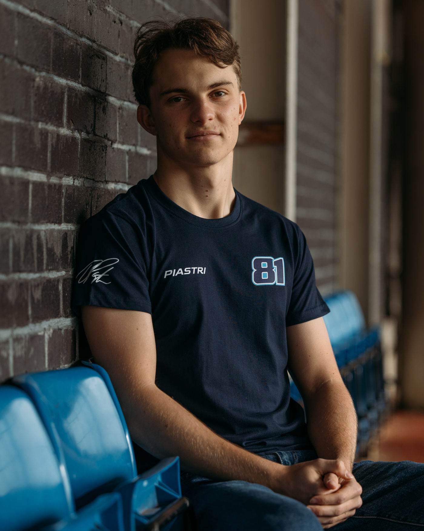 Man wearing Piastri 81 t-shirt with race number, last name, and signature, sitting on blue seats, showcasing casual sportswear fashion.