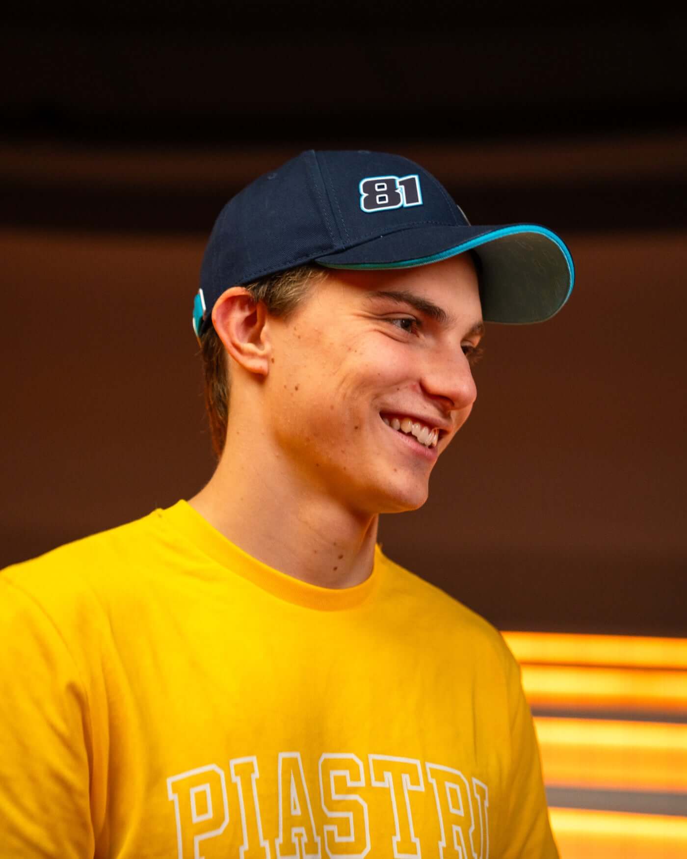 Young man wearing navy Signature Cap with race number 81 and yellow shirt, smiling in casual setting.