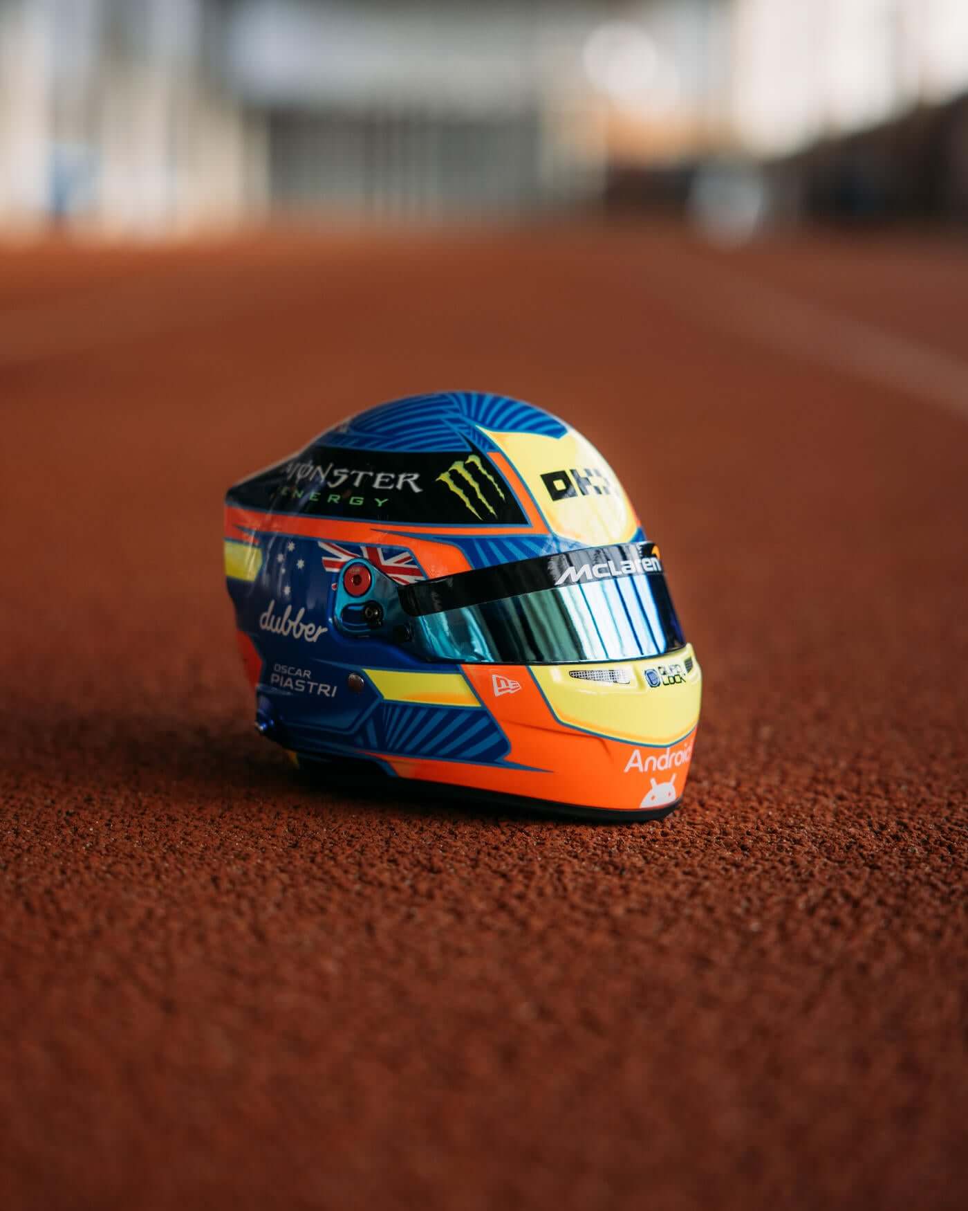 1:2 scale replica of Oscar Piastri's 2024 season helmet with red, blue, and yellow design placed on a track.