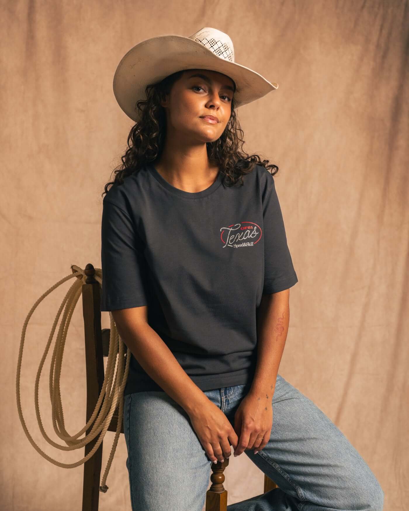 Woman wearing Texas 2024 T-Shirt in Asphalt, inspired by Circuit of the Americas and Western culture, seated with cowboy hat and rope.