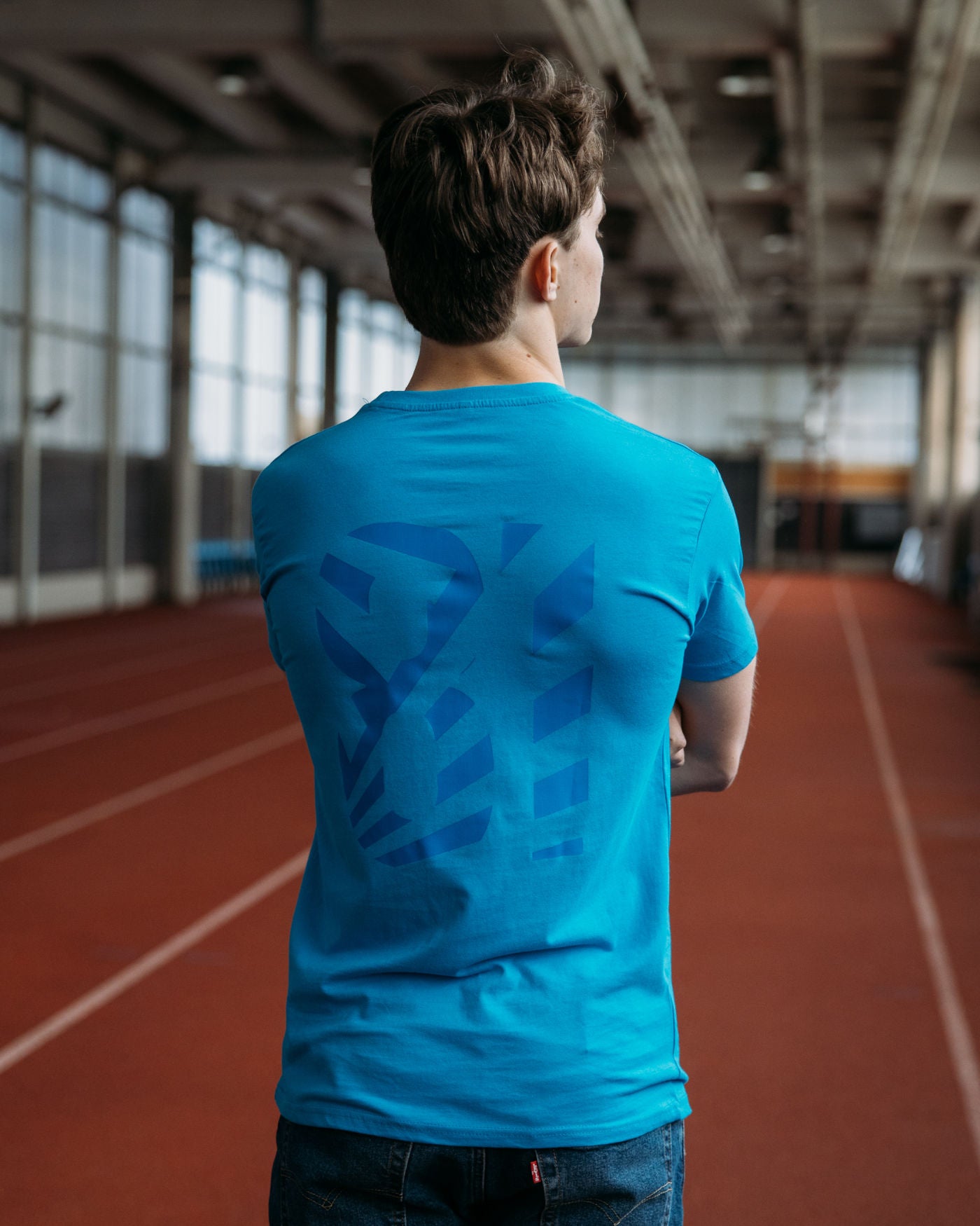 Man wearing Keep Your Cool T-Shirt in Aqua with OP logo and dazzle effect on the back, standing on an indoor running track.