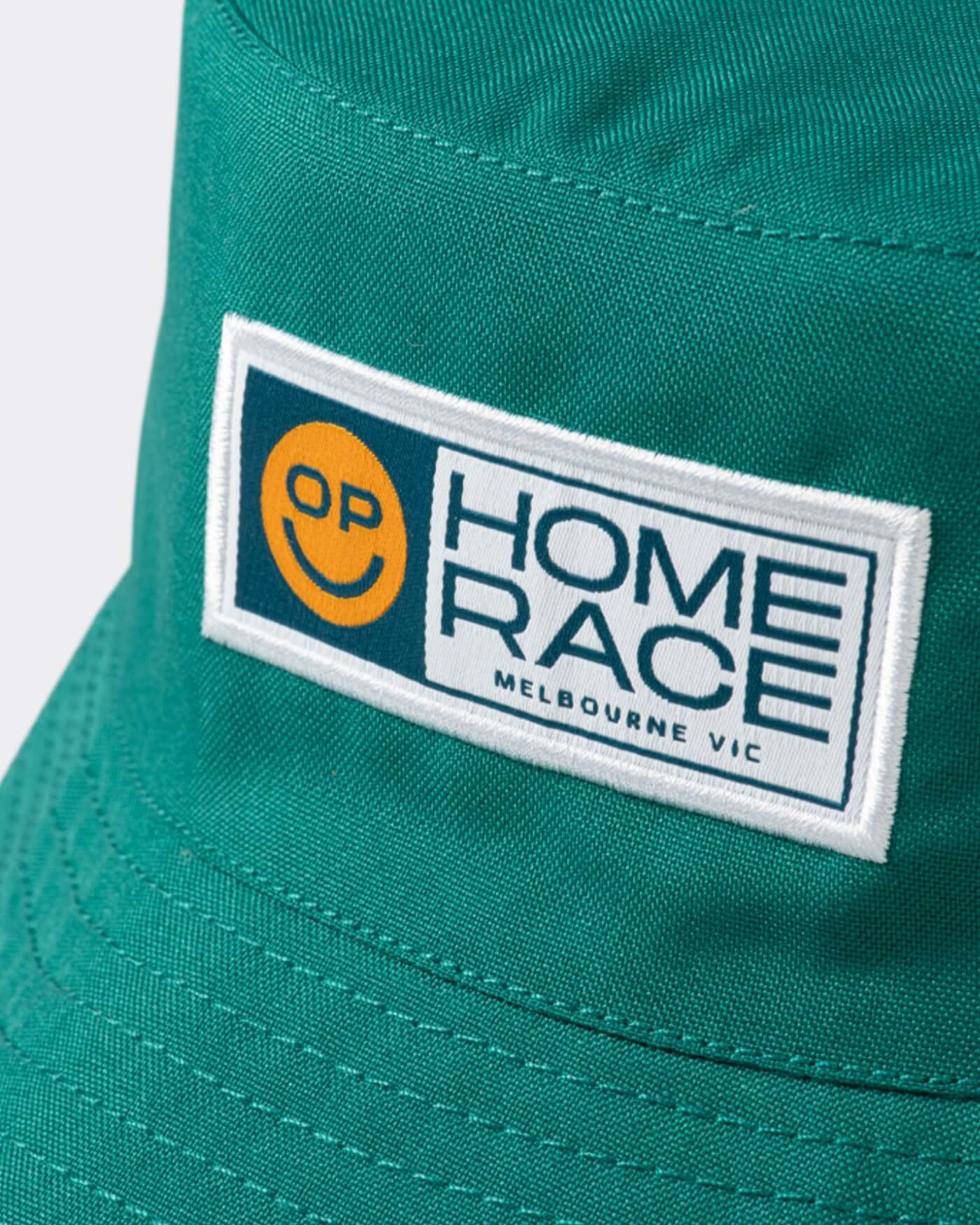 Close-up of the Home Race '26 Bucket Hat showcasing the OP smiley design in green and gold fabric.