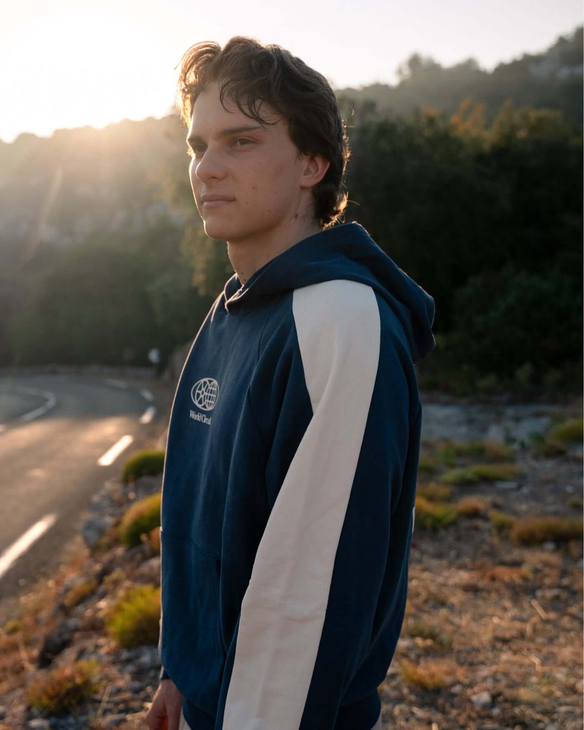 Model wearing World Circuit Hoodie - Blue with off-white sleeves in a scenic outdoor setting.