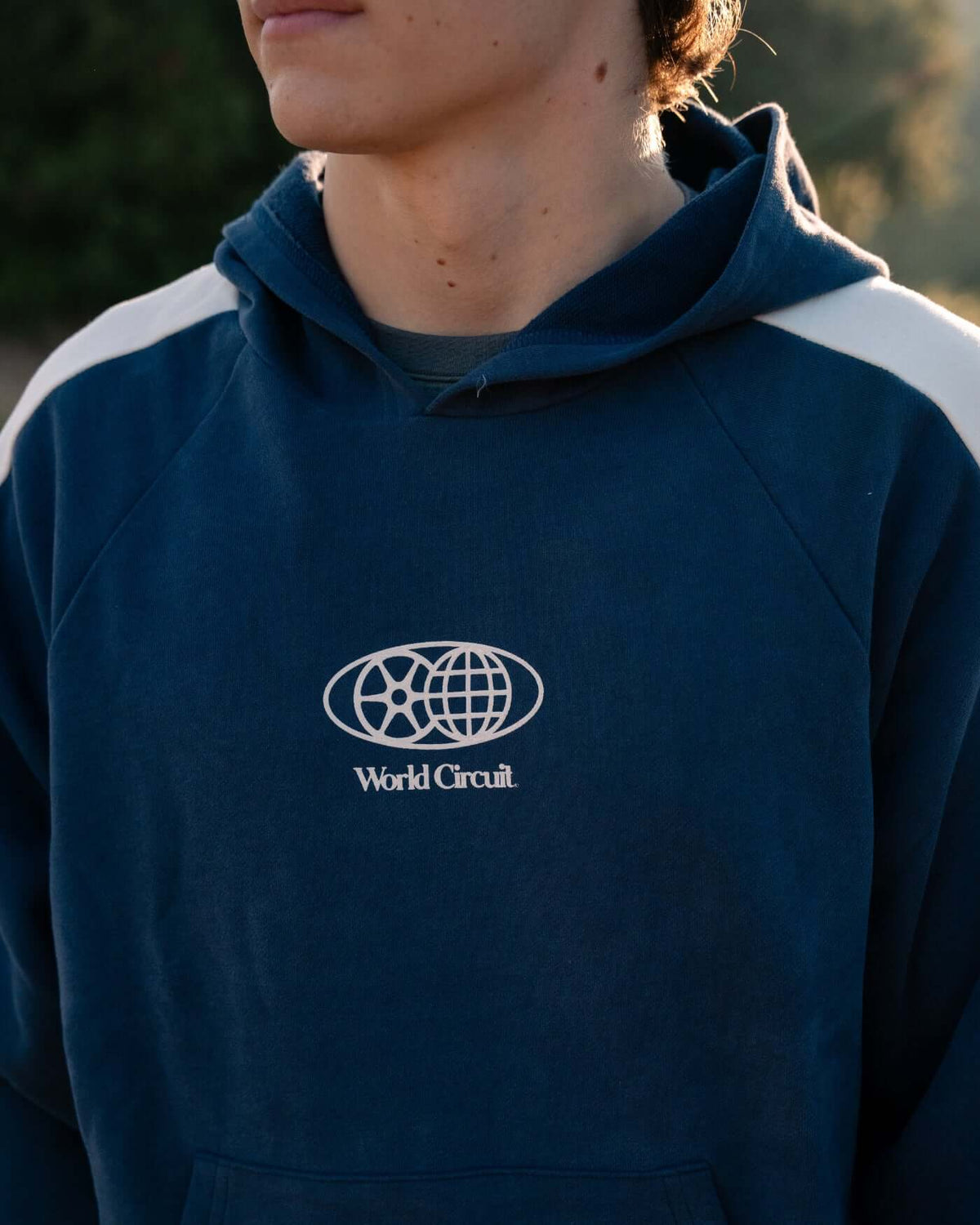 Person wearing a World Circuit Hoodie - Blue with globe emblem and off-white racing panels.