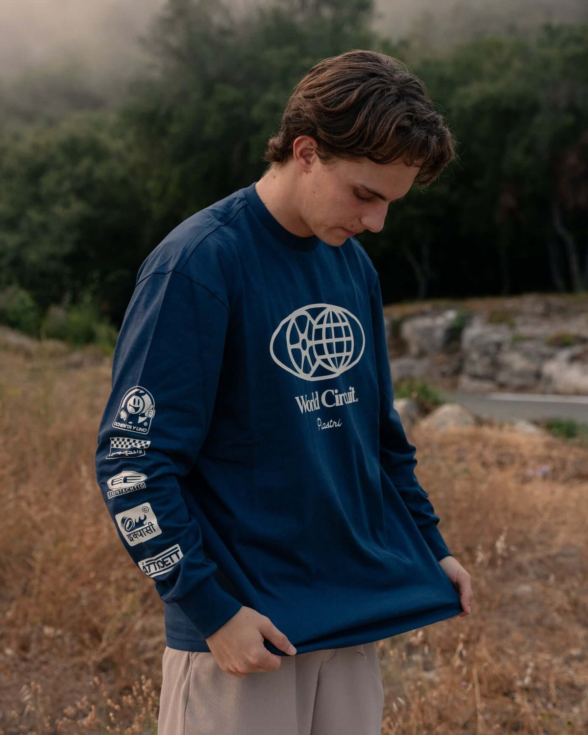 Model wearing World Circuit Long Sleeve T-Shirt - Blue with globe graphic and sponsor logos on sleeves.