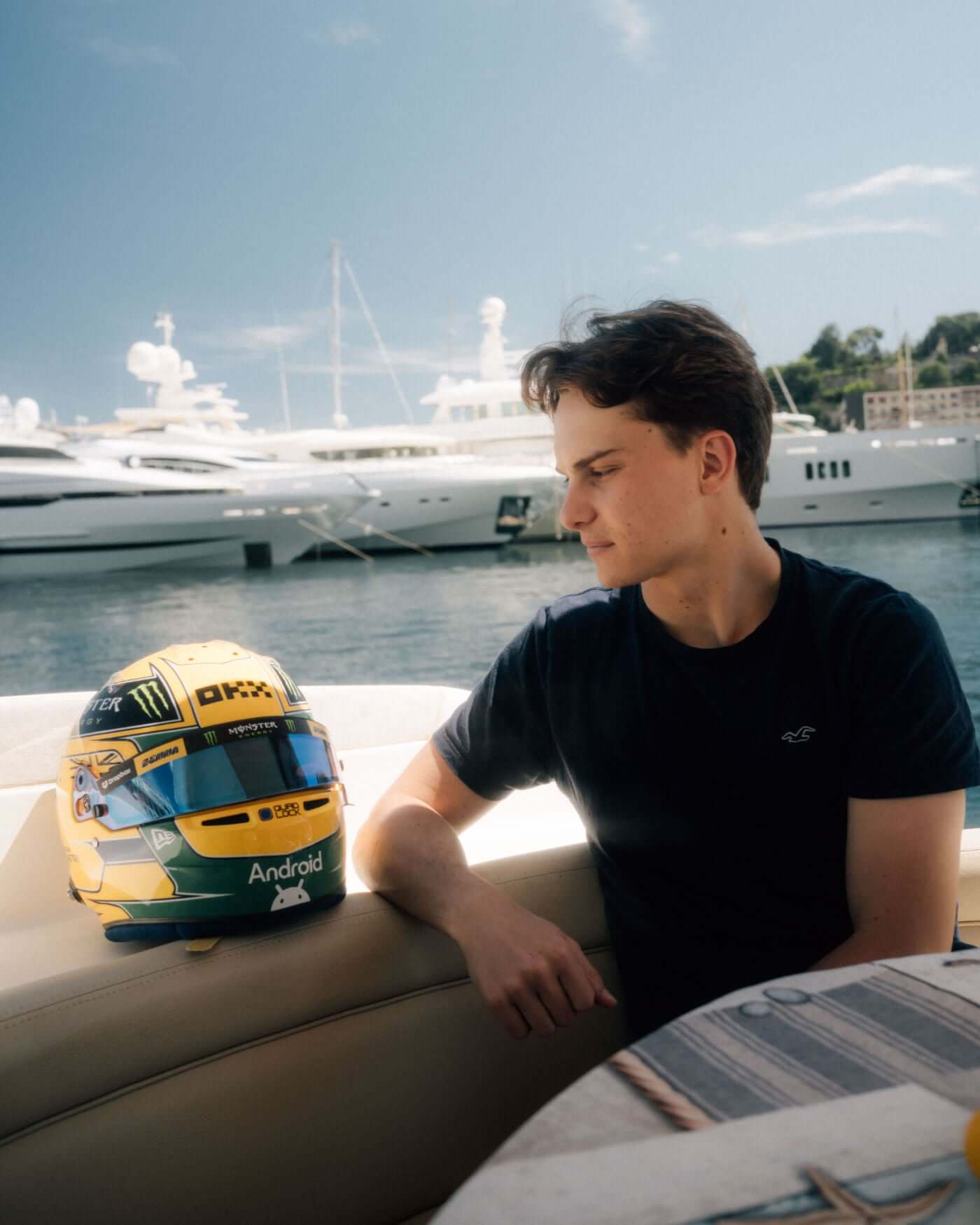 Oscar Piastri sitting beside 1:2 scale Monaco 2024 Ayrton Senna tribute helmet with luxury yachts in the background.